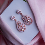 A pair of rose gold bejeweled statement earrings with a sparkling design, displayed on a pink surface with a white flower and a pale pink card.